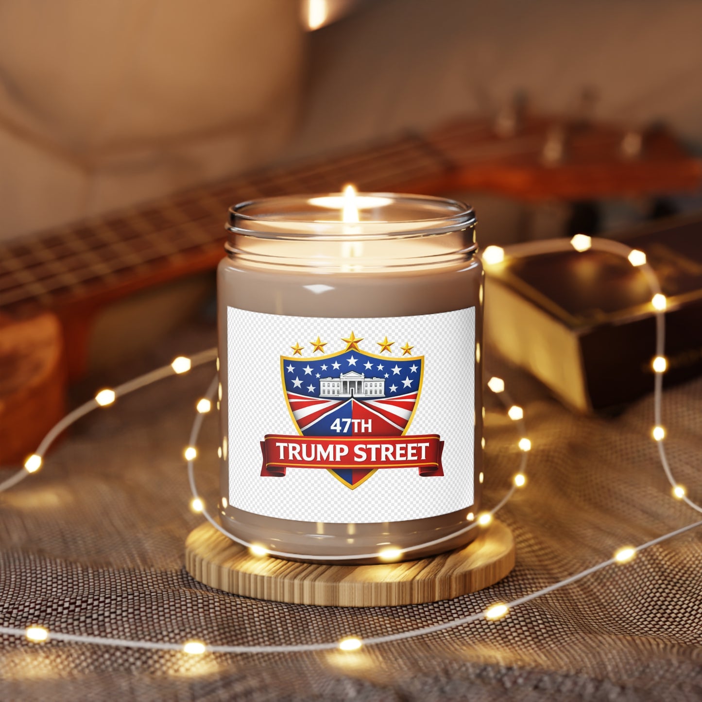 Candle in a jar with '47th Trump Street' label on a wooden coaster with fairy lights, guitar in the background